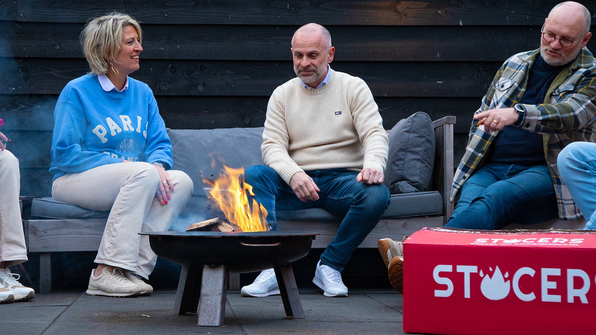 Groep mensen die rond een buitenvuur zitten met een STOCERS-doos aanmaakhout; sfeerbeeld voor haardhout en aanmaakproducten.